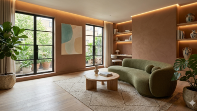 "تصميم غرفة معيشة عصرية تعتمد أحدث صيحات ديكور 2026، تضم أريكة منحنية باللون الأخضر الزيتوني، جدران ترابية دافئة، وطاولة خشبية مستديرة مع إضاءة خفيفة ونباتات طبيعية داخلية."