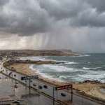"صورة القمر الصناعي توضح سحب الرعد الكثيفة Sheets of Rain وهي تتساقط فوق البحر المتوسط وتغطي الطريق الساحلي الدولي المبتل الممتد من الإسكندرية نحو شمال سيناء، مع لوحات إرشادية رقمية تحذر من انعدام الرؤية والسيول 2026، كإشارة لبدء سقوط الأمطار."
