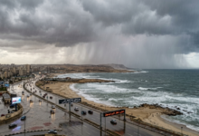 "صورة القمر الصناعي توضح سحب الرعد الكثيفة Sheets of Rain وهي تتساقط فوق البحر المتوسط وتغطي الطريق الساحلي الدولي المبتل الممتد من الإسكندرية نحو شمال سيناء، مع لوحات إرشادية رقمية تحذر من انعدام الرؤية والسيول 2026، كإشارة لبدء سقوط الأمطار."