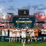 "احتفالات لاعبي نادي الزمالك على أرضية الملعب مع الجماهير البيضاء بعد تصدر ترتيب دوري نايل، ويظهر في الخلفية لوحة النتائج مع جدول الترتيب الذي يظهر الزمالك في المركز الأول."