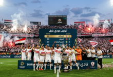 "احتفالات لاعبي نادي الزمالك على أرضية الملعب مع الجماهير البيضاء بعد تصدر ترتيب دوري نايل، ويظهر في الخلفية لوحة النتائج مع جدول الترتيب الذي يظهر الزمالك في المركز الأول."