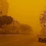 "مشهد حي لعاصفة ترابية تجتاح سوهاج الآن، حيث تغطي سحب الرمال الصفراء الكثيفة المباني والشوارع وتؤدي لانعدام الرؤية على الطرق السريعة، مع ظهور سيارة إسعاف ترمز لحالة الطوارئ المعلنة في المحافظة."