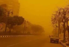 "مشهد حي لعاصفة ترابية تجتاح سوهاج الآن، حيث تغطي سحب الرمال الصفراء الكثيفة المباني والشوارع وتؤدي لانعدام الرؤية على الطرق السريعة، مع ظهور سيارة إسعاف ترمز لحالة الطوارئ المعلنة في المحافظة."